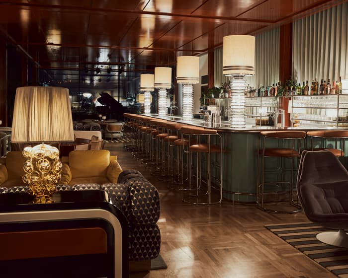 Stylish bar with a long counter lined with high stools, large lamps, and a variety of drinks behind. Chic lounge furniture, warm ambient lighting, and a grand piano in the background.