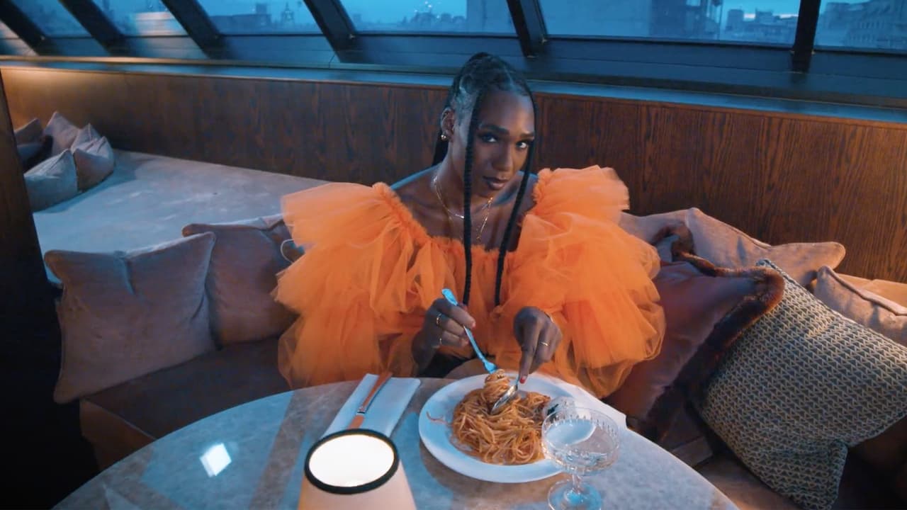 Person in an orange tulle dress eating spaghetti at a candlelit table inside a cozy, glass-roofed lounge with cityscape views. Velvet pillows surround the seating area.