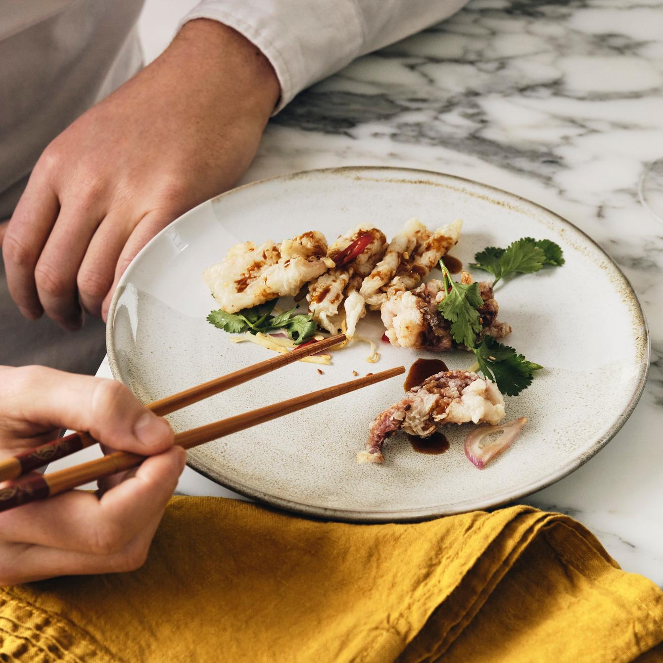 A person uses chopsticks to pick up a piece of fried food on a white plate garnished with greens, set on a marble table with a yellow napkin and a glass of white wine.