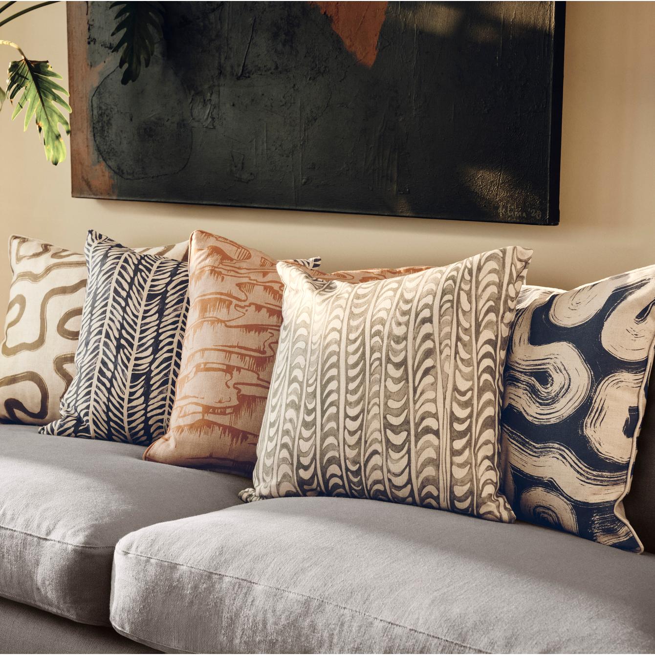 Five patterned cushions rest on a grey sofa, with a green potted plant and abstract wall art in the background.