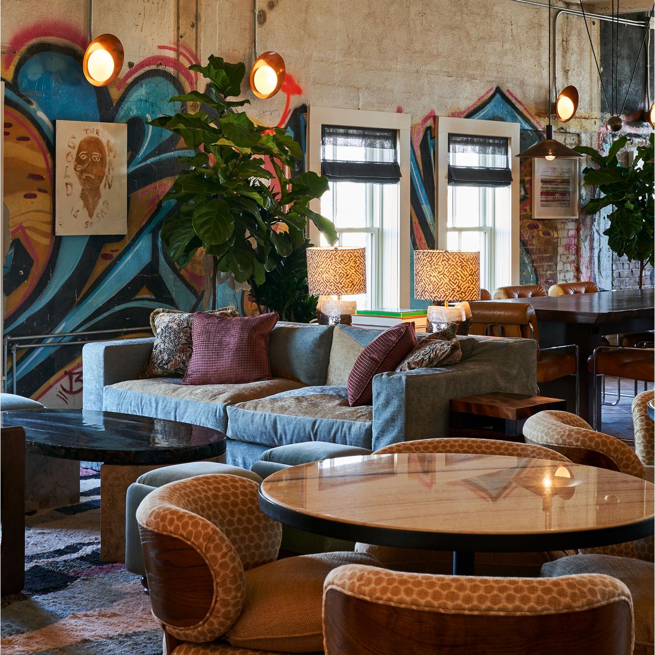 A cozy seating area with a gray sofa, patterned chairs, and round tables is in a room featuring large windows, colorful graffiti walls, hanging lights, and indoor plants.