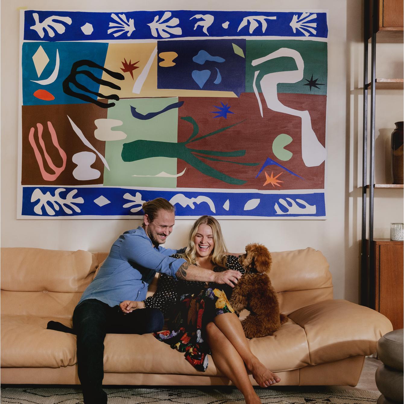A couple on a beige sofa joyfully plays with a small brown dog in a living room adorned with a colorful abstract painting, a wooden shelf with ceramics, and candles.