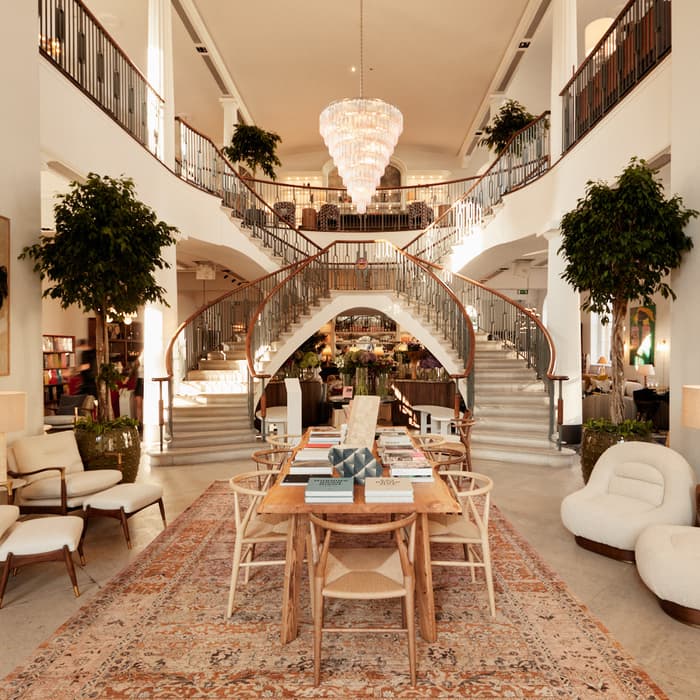Elegant lobby with a central wooden table holding books, surrounded by white armchairs and lush plants; a grand staircase leads to an upper level adorned with a crystal chandelier and artwork.