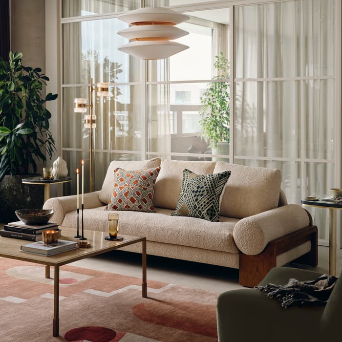 A contemporary living room featuring a beige sofa with patterned cushions, a coffee table with candles and books, adjacent to armchairs, houseplants, and modern decor, illuminated by natural light and an overhead lamp.