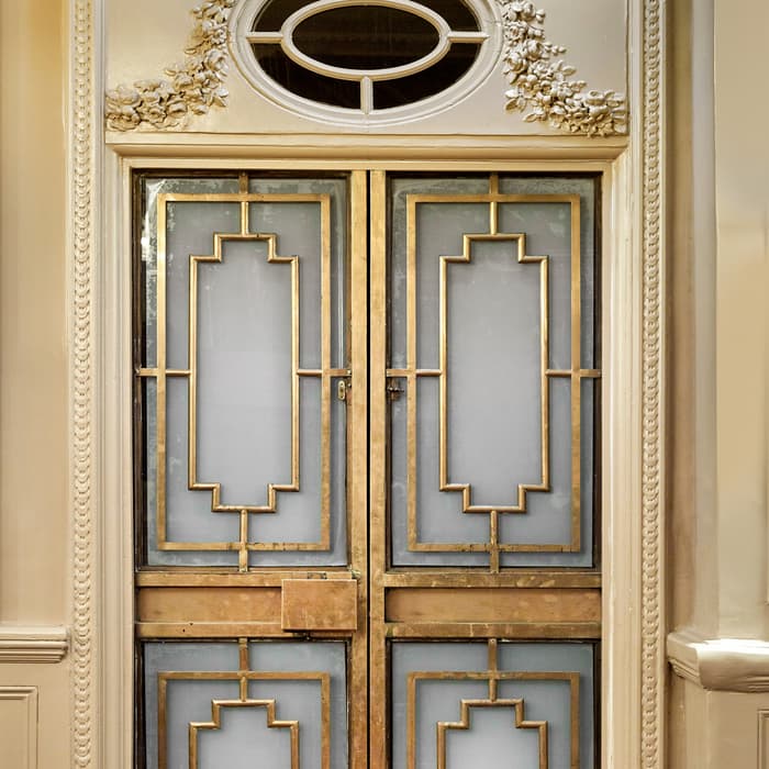 An ornate set of double doors, featuring gold geometric frames and frosted glass panes, situated within an elegant, classical interior with detailed molding and a small, decorative oval window above.