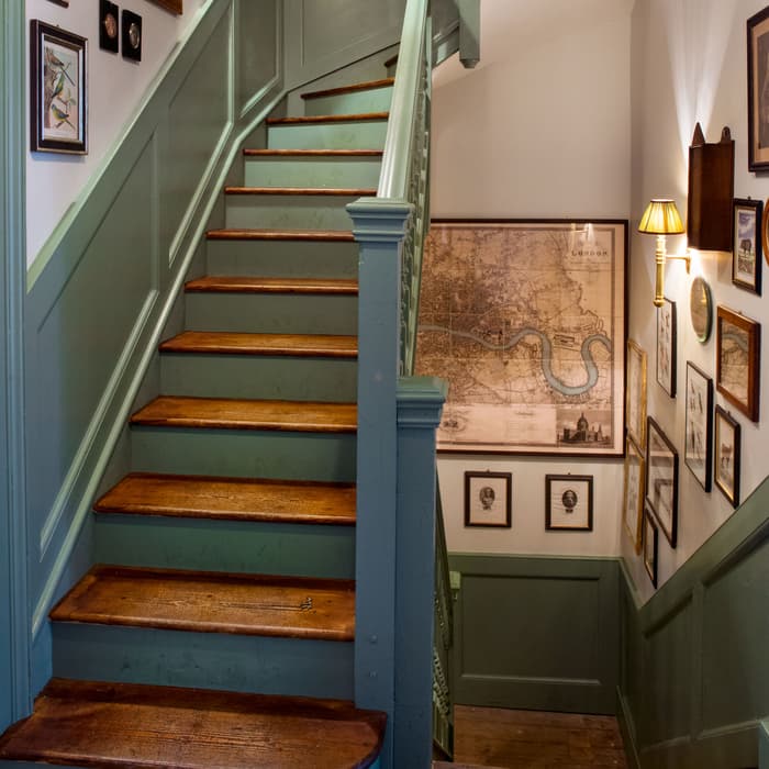 A staircase with light green railings and wooden steps ascends in a hallway. The walls are adorned with framed maps, drawings, and photographs, illuminated by wall-mounted lamps.