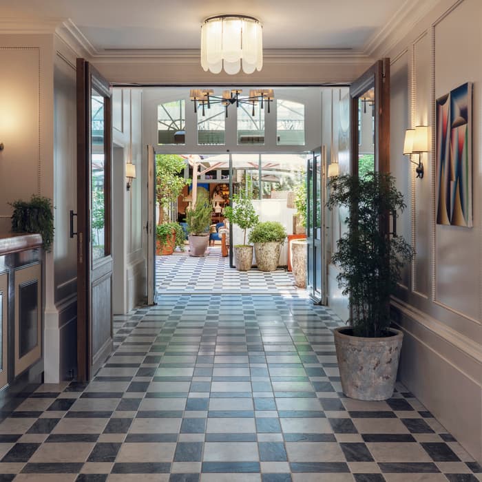 Elegant hallway features black-and-white checkered flooring, plants, and wall-mounted lamps; open double doors lead to an outdoor garden area with potted plants, seen through glass-panel doors.