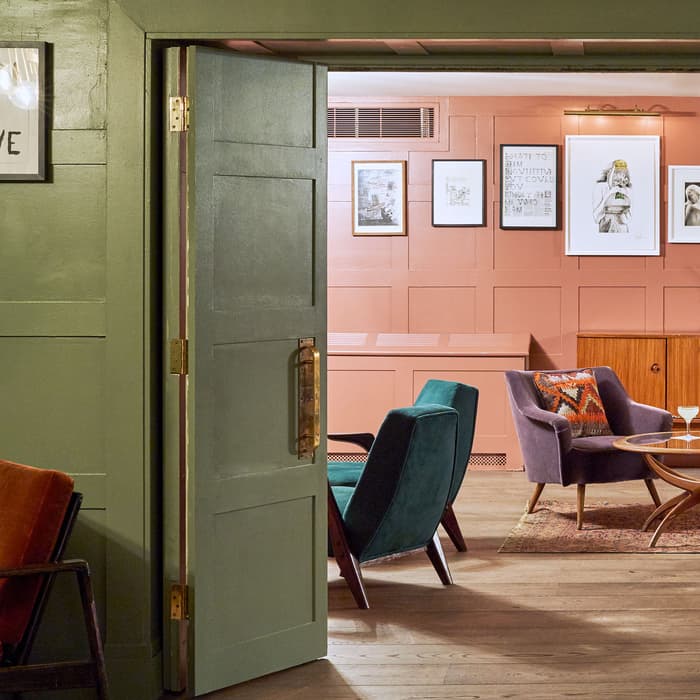 A partially open green door reveals a cozy sitting area with colorful chairs, a round table with drinks, and a wall with framed artworks in a room with warm lighting.