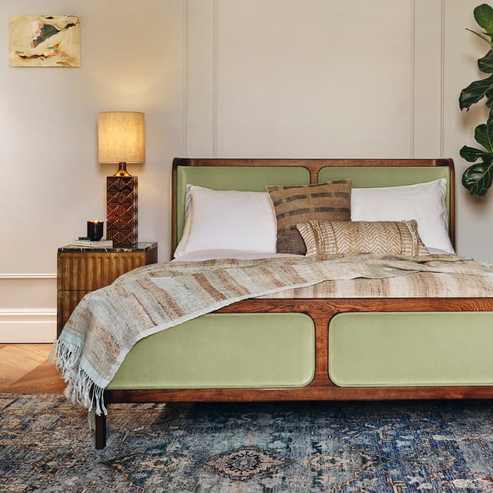 Bed with green upholstered headboard, adorned with patterned pillows and a woven throw, situated in a cozy bedroom featuring a wooden nightstand, decorative lamp, wall art, and a blue patterned rug.