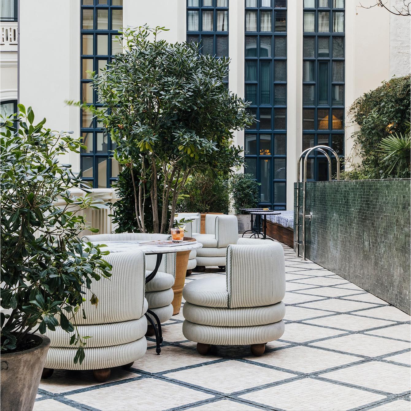 Plush seats surround a table with drinks in a stylish outdoor patio adorned with potted plants and framed by tall windows and a green-tiled wall.