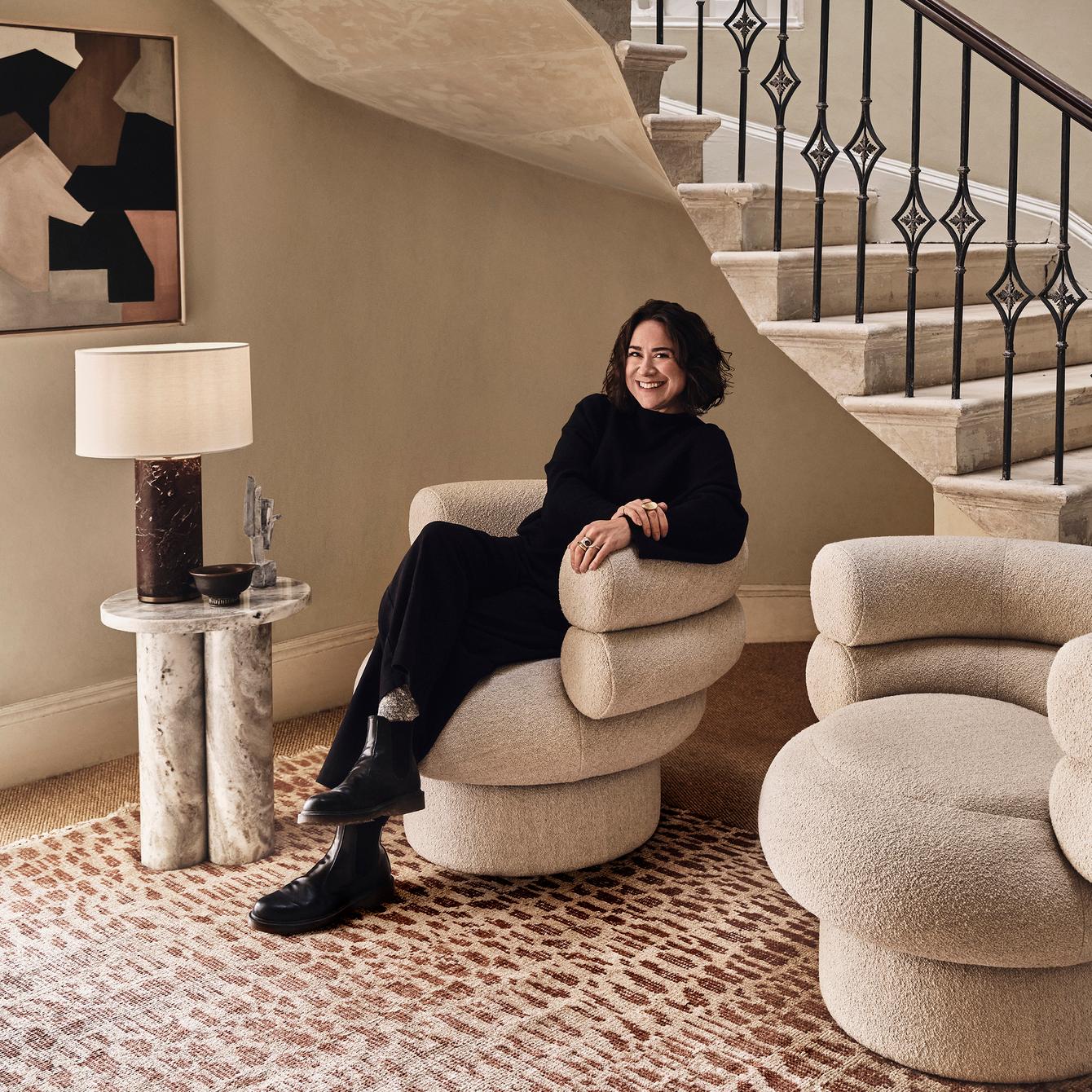 A person in black clothing sits smiling on a cushioned armchair beside a lamp table, under a staircase with an ornate railing. The room features a rug and a minimalist decor.