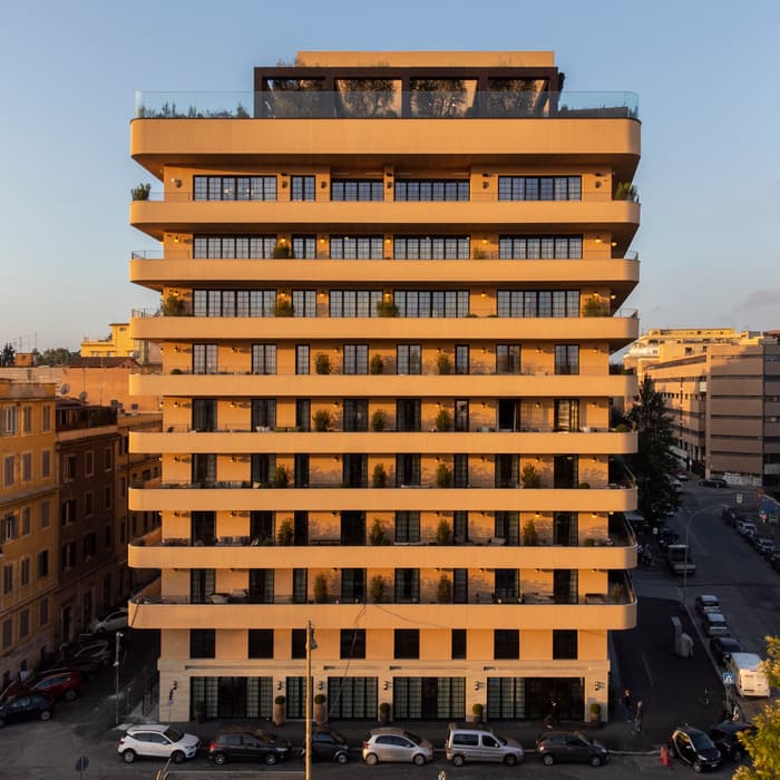 A multi-story building with wide balconies and large windows, exhibiting modern architecture at sunset, situated in an urban area with parked cars and adjacent residential buildings.