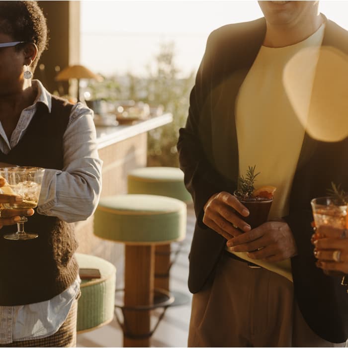 Three people hold drinks and engage in conversation, illuminated by warm, natural light. They are standing in a stylish outdoor lounge with bar stools and greenery in the background.