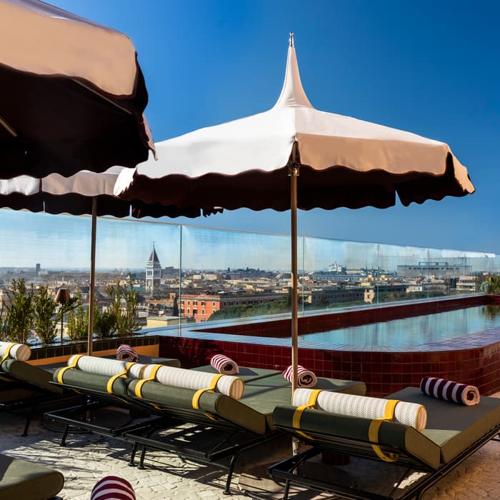 Sun loungers with striped towels under large umbrellas face a rooftop pool, which is surrounded by a glass barrier overlooking a cityscape on a sunny day.