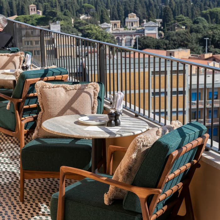 A small round table set with napkins and condiments is flanked by green cushioned wooden chairs on a tiled balcony overlooking a cityscape with trees and distant buildings.