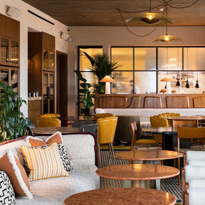 Comfortable seating area with couches and pillows, tables arranged, and modern pendant lights overhead; situated in a stylish, well-lit lounge featuring a mix of wooden and lush green plant decor.