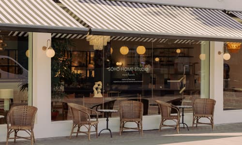 A chic storefront with striped awnings features "SOHO HOME STUDIO" text on the window. Wicker chairs and tables rest on the sidewalk, in front of an inviting, warmly-lit interior.