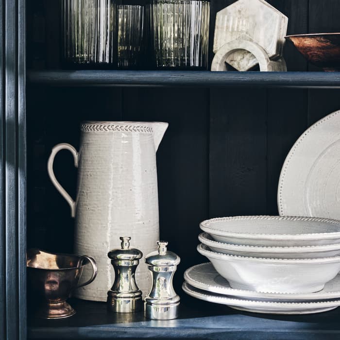 Glassware and ceramics sit arranged on a dark wooden shelf, featuring glasses, a white pitcher, silver salt and pepper shakers, a stack of plates, and small silver and copper vessels.