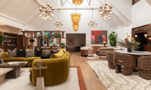 A large living room features a mustard-yellow velvet couch with patterned cushions, surrounded by modern decor, artwork, plants, wooden furniture, and hanging light fixtures under a vaulted, white ceiling.