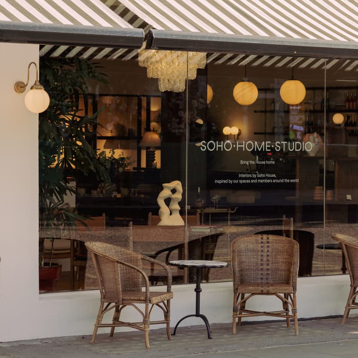 Wicker chairs and small tables sit outside a shop with striped awnings. The glass displays a sign reading: "SOHO HOME STUDIO. Bring the house home. inspired by Soho House." Inside, warm lighting and decor are visible.