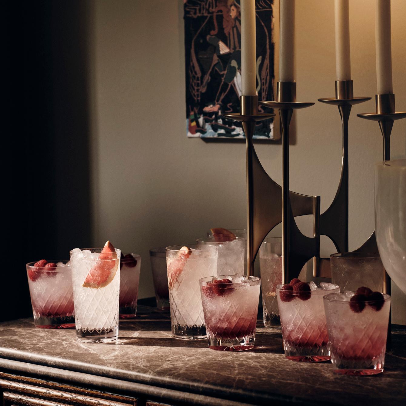 Several glasses filled with ice, raspberries, and a pink liquid rest on a marble-topped table. Behind them, a brass candelabra with white candles and a framed artwork adorn the wall.