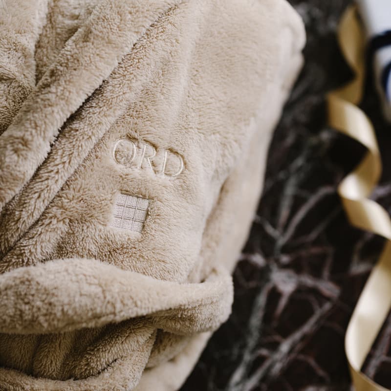 A folded beige bathrobe with "ORD" and a small square embroidered on it lies on a dark patterned surface, next to ribbon and other blurred objects. "SOHO HOUSE" is visible on the label.