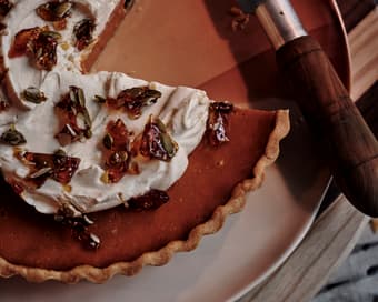 A partially-sliced pumpkin pie with whipped topping and brittle pieces, resting on a striped cutting board, beside a knife on a red plate in a cozy, homey setting.