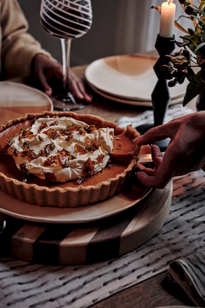 A hand places a pumpkin pie topped with whipped cream on a table set for a cozy meal, surrounded by plates, a wine glass, and a candle.