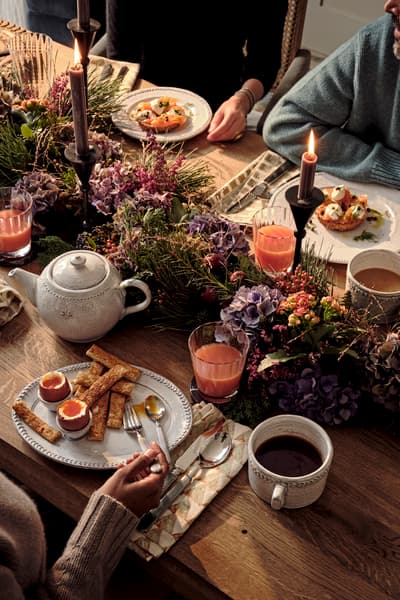 A cozy breakfast setting with three people around a wooden table, enjoying poached eggs, toast, tea, and juice. The table is decorated with flowers, candles, and rustic crockery.
