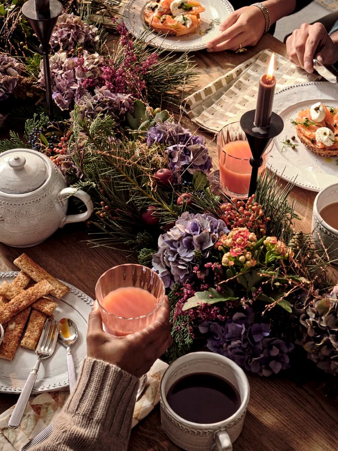 Table set for brunch with tea, juice, soft-boiled eggs, bread, and greenery centerpiece. People using utensils eating in a cozy, warmly lit environment. Plates and cups neatly arranged.