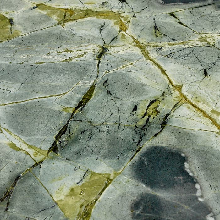 A slab of greenish-gray marble with a network of cracks and yellowish veins runs throughout. The surface appears polished, displaying a variety of darker and lighter patches.
