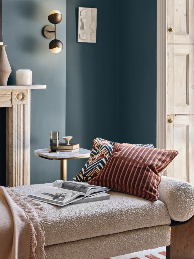A chaise lounge with pillows and a blanket sits beside an open book and table with a glass, within a cozy room featuring a fireplace, wall art, and an open window with greenery outside.