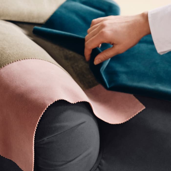 A hand is placing fabric swatches in olive green, pink, and blue on the armrest of a dark gray sofa, suggesting a selection process for upholstery.