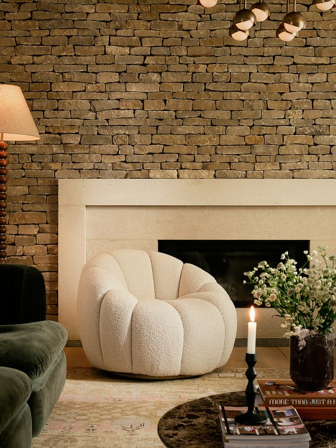 A living room features a green velvet sofa with cushions, two white plush armchairs, a stone fireplace, a standing lamp, and a marble coffee table with a candle, books, and a vase of flowers.