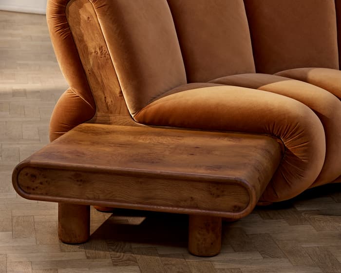 A plush, curved brown couch with wooden armrests is situated on a wooden floor in a white, minimally-decorated room.