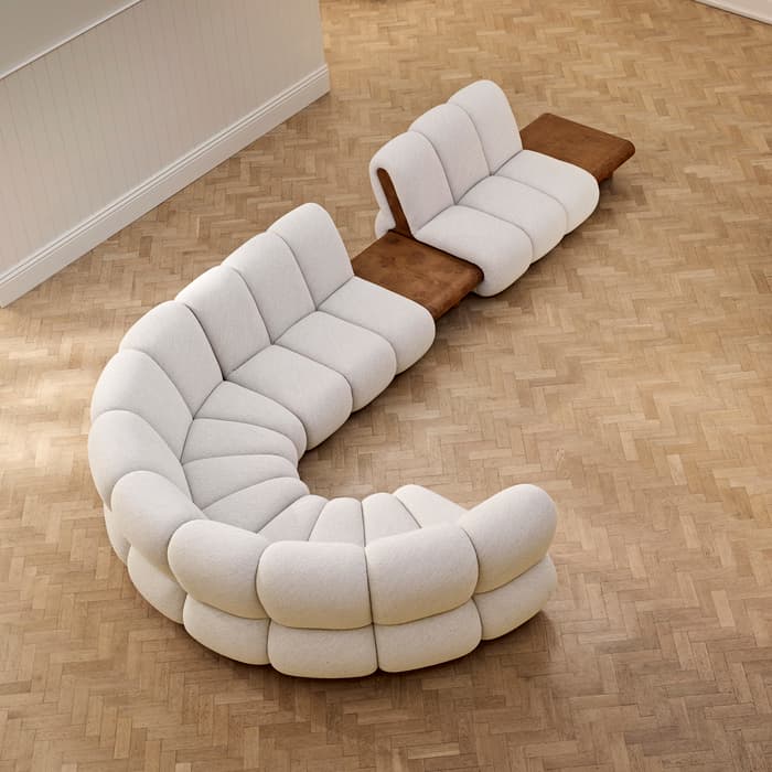 Curved, beige modular sofa with brown wooden side tables placed on a light wooden herringbone floor. Another matching, smaller sofa is located in the back corner of the room.