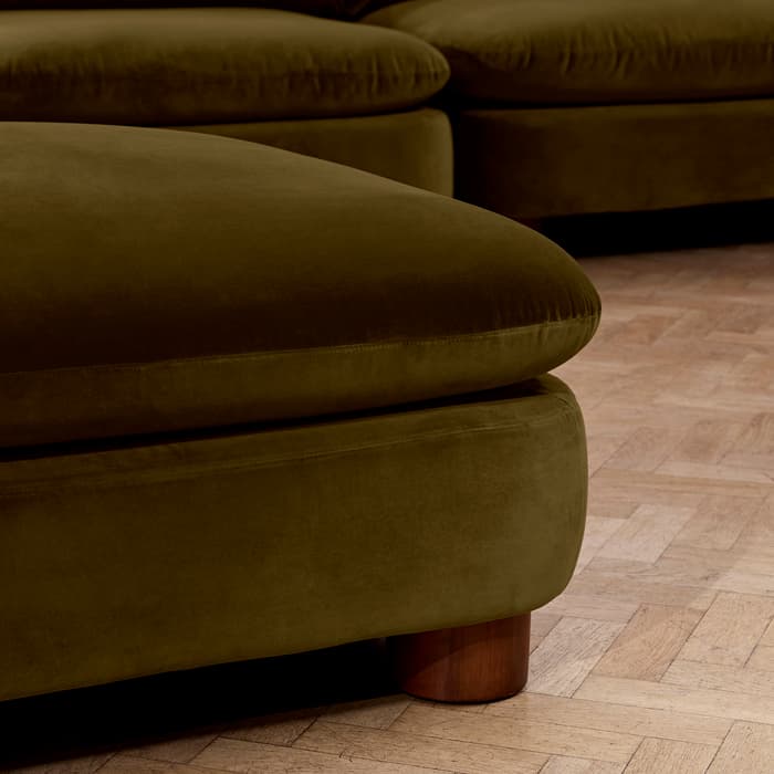 A green velvet sofa with rounded wooden legs is positioned on a polished herringbone-patterned wooden floor in an indoor setting.
