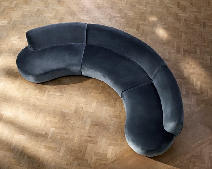 A curved, gray sofa sits empty on a light herringbone wood floor, viewed from above, with no other furniture or decor visible in the room.