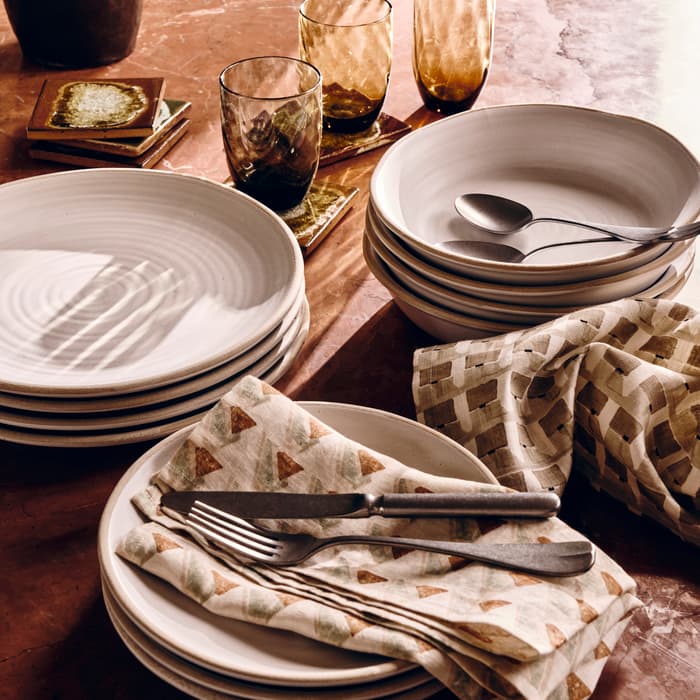 Stacked white plates and bowls with cutlery and patterned napkins arranged on a marble table, surrounded by amber-colored glasses, ceramic coasters, a vase, and a patterned chair in the background.