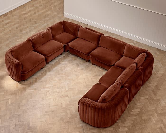 A large, brown, U-shaped sectional sofa is situated on a light wooden parquet floor in a neutral-colored room with white paneled walls.