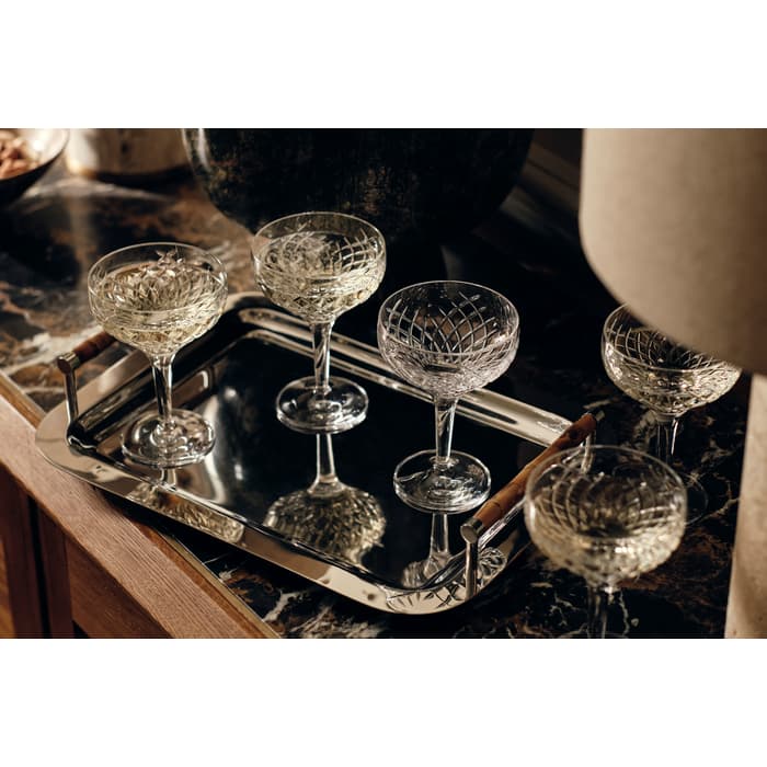 Five crystal glasses with intricate patterns rest on a polished silver tray. Each glass holds a light-colored liquid. The tray is placed on a dark marble surface with various decorative items around.