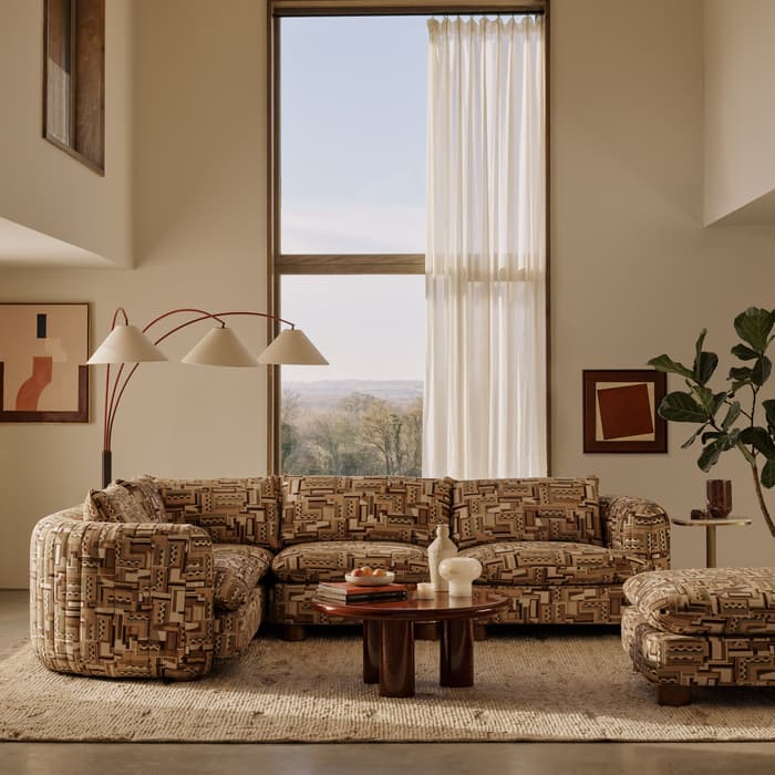 Patterned sectional sofa placed in a spacious living room; a large window with sheer curtains behind it, flanked by artwork, a geometric floor lamp, a coffee table with decor, and a rug.