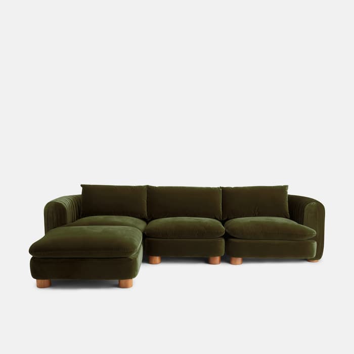 Green velvet sectional sofa with cylindrical wooden legs, featuring a chaise on the left. Positioned against a plain white background.
