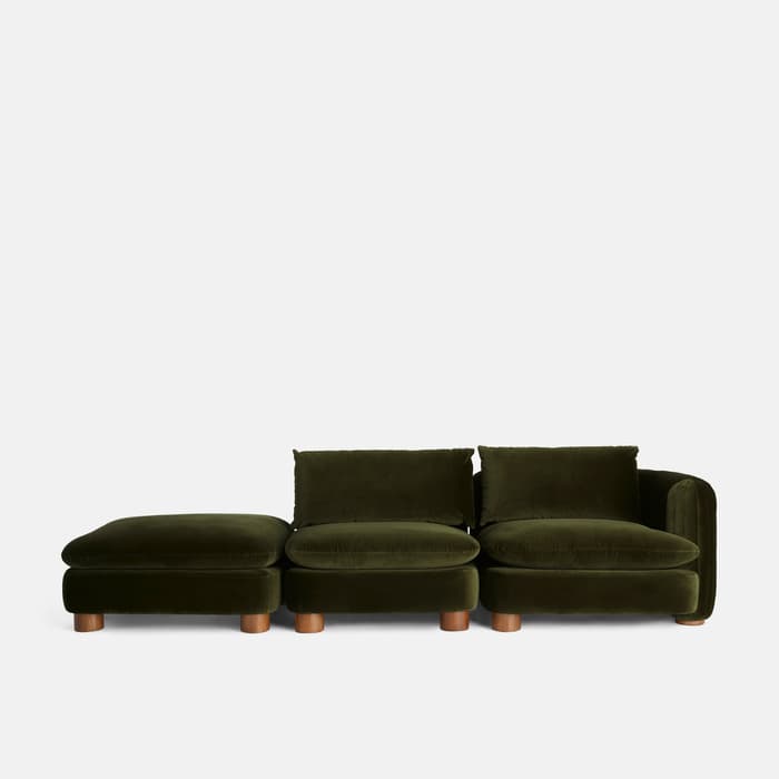 Green velvet modular sofa with three sections including a chaise lounge, positioned against a plain white background. The seating rests on cylindrical wooden legs. The sofa has a modern, minimalist design.