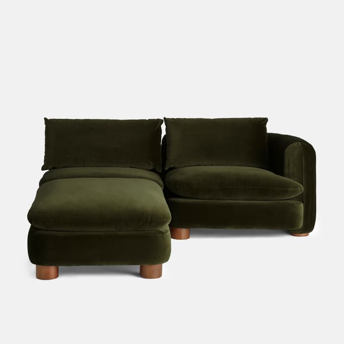 A dark green velvet sectional sofa with two large back pillows rests against a white background. The left portion serves as a chaise lounge with wooden legs.