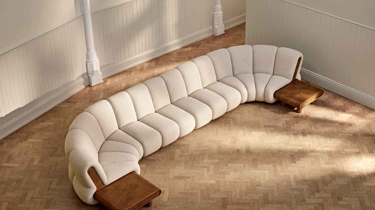 Curved, modern beige sofa with cushioned segments; placed in an empty room with light wood herringbone flooring, ornate white pillars, and cream-colored wainscoting on the walls.