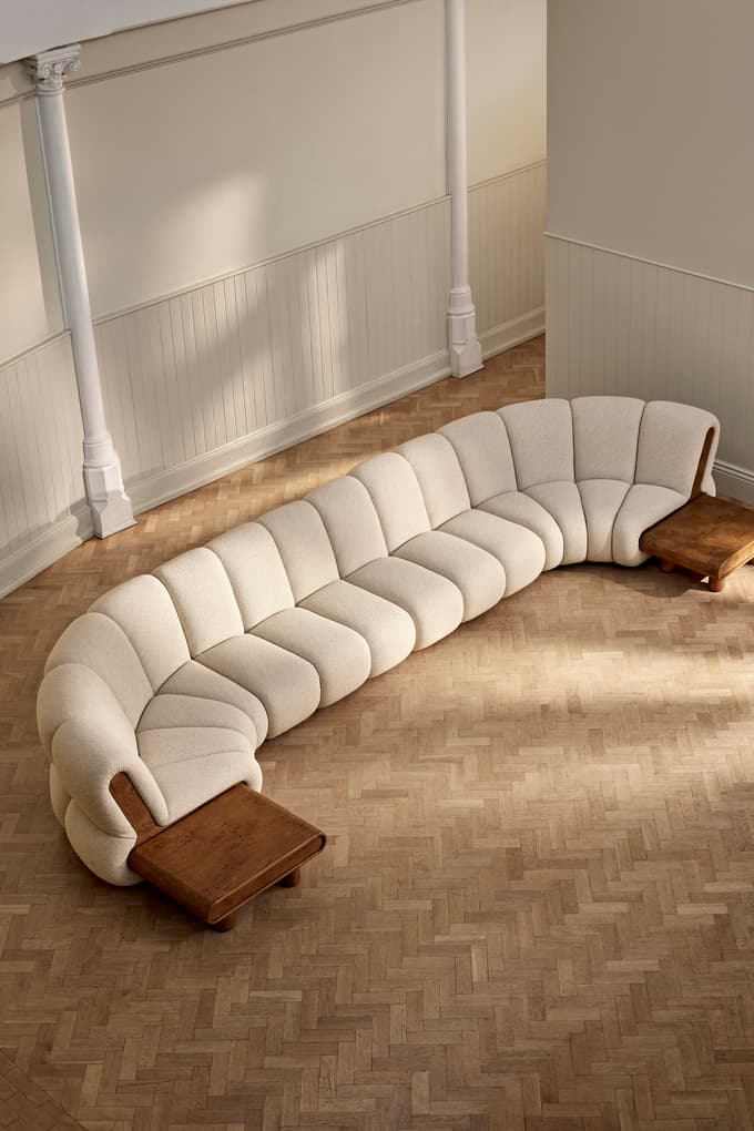 A large, beige, curved sectional sofa with built-in wooden side tables stands on a herringbone-patterned wooden floor in a spacious room with cream walls and white pillars.