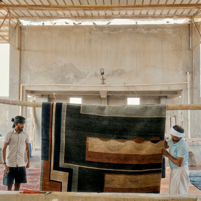Two men hang a large geometric-patterned rug on a wooden beam, inside a spacious, industrial-looking room with a high, metal-framed roof.