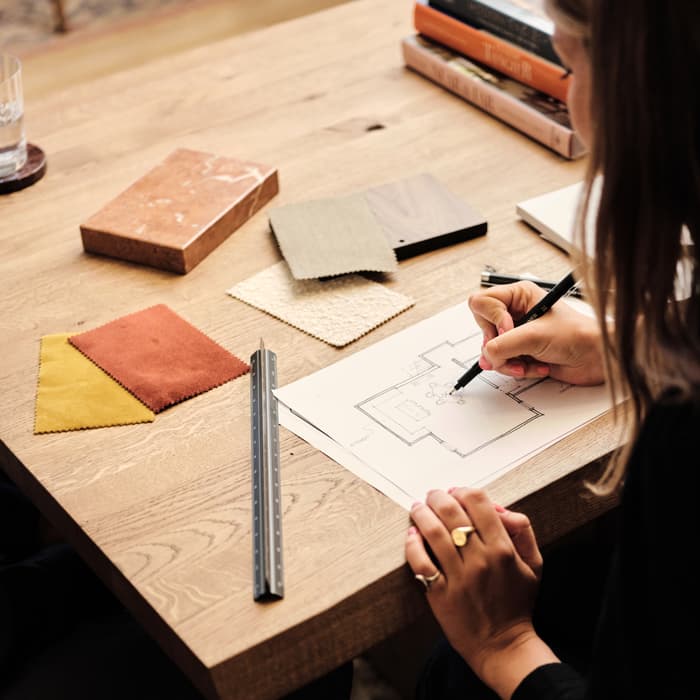 A person draws a floor plan on paper at a wooden table with fabric swatches, a ruler, a block, and stacked books nearby in a cozy, indoor setting.