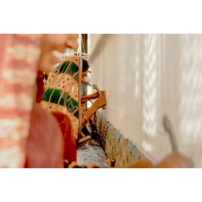 Two women, dressed in colorful, patterned fabrics, meticulously tie knots on a large, vertical tapestry loom in a sunlit room. One woman's face is blurred in the foreground.